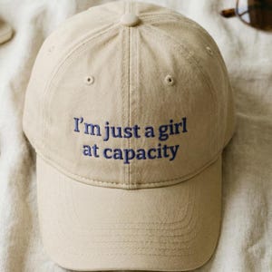 May include: A beige baseball cap with the embroidered phrase "I'm just a girl at capacity" in blue thread. The cap has a curved brim and a button on top. The background is a neutral fabric.