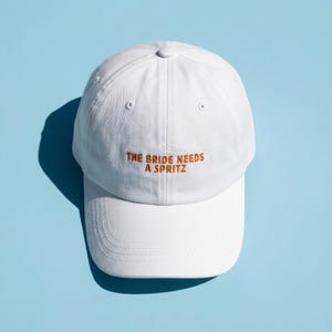 Puede incluir: Gorra de béisbol blanca con visera curva. La gorra presenta el texto bordado "THE BRIDE NEEDS A SPRITZ" en color marrón. El sombrero está sobre un fondo azul claro.