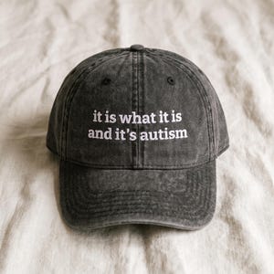 May include: A charcoal grey baseball cap with a washed, distressed finish. The front panel features white embroidered text that reads, "it is what it is and it's autism." The cap has a curved brim and a structured crown with a top button.