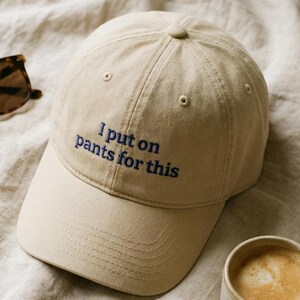 May include: A beige baseball cap with the embroidered phrase "I put on pants for this" in blue. The cap is made of a soft fabric and has a curved brim. A cup of coffee is visible in the bottom right corner.