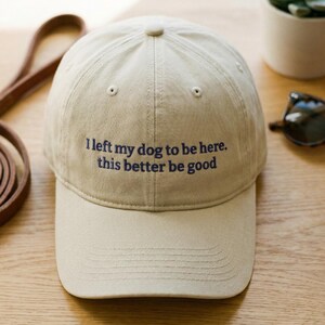 May include: A tan baseball cap with the embroidered text "I left my dog to be here. this better be good" in navy blue. The cap has a curved brim and a button on top. A leather dog leash is visible in the background.