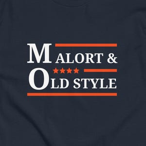 May include: A navy blue t-shirt featuring the text "MALORT & OLD STYLE" in white and orange. The text is arranged in two lines with orange horizontal lines above and below the words "OLD STYLE". The shirt has a crew neck and short sleeves.