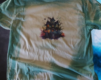 May include: A light green t-shirt featuring a haunted house graphic. The graphic is a detailed illustration of a spooky mansion with trees and bats. The shirt has a bleached design, creating a unique, distressed look. The t-shirt is a crew neck.