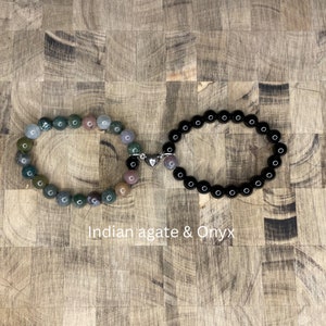 Peut inclure: Deux bracelets reli&eacute;s par un charm en forme de c&oelig;ur argent&eacute;. Un bracelet est compos&eacute; de perles d'onyx noir et l'autre de perles d'agate indienne multicolores. Les bracelets sont sur une surface en bois.