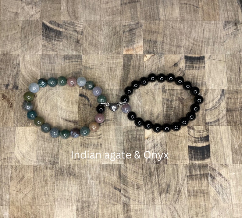 Peut inclure: Deux bracelets reli&eacute;s par un charm en forme de c&oelig;ur argent&eacute;. Un bracelet est compos&eacute; de perles d'onyx noir et l'autre de perles d'agate indienne multicolores. Les bracelets sont sur une surface en bois.