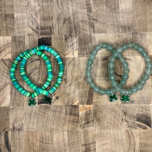 May include: Two sets of green and blue beaded bracelets with shamrock charms. The bracelets are on a wooden surface.