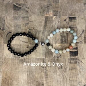 Peut inclure: Deux bracelets en perles reli&eacute;s par un charm en forme de c&oelig;ur argent&eacute;. Un bracelet est en onyx noir et l'autre est un m&eacute;lange de perles d'amazonite bleu clair, vert et brun. Les bracelets sont sur une surface en bois. Amazonite & Onyx