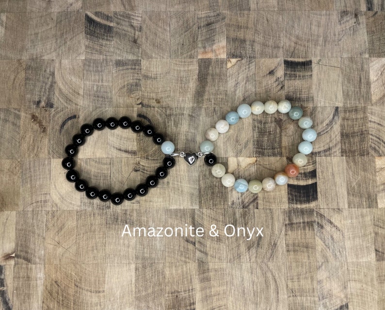 Peut inclure: Deux bracelets en perles reli&eacute;s par un charm en forme de c&oelig;ur argent&eacute;. Un bracelet est en onyx noir et l'autre est un m&eacute;lange de perles d'amazonite bleu clair, vert et brun. Les bracelets sont sur une surface en bois. Amazonite & Onyx
