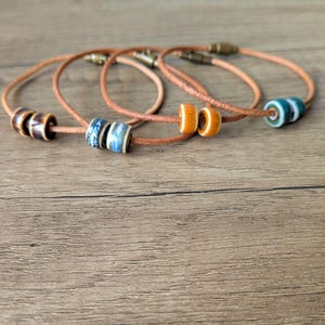 May include: Four leather bracelets with ceramic beads in various colors. The beads include brown, blue, orange, and teal. The bracelets have a simple design with a clasp closure, and are displayed on a wooden surface.