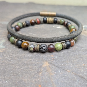 May include: Two bracelets. One has a dark grey leather band with a gold-coloured clasp. The other is made of various coloured beads, including green, red, and black. The bracelets are displayed on a textured grey surface.