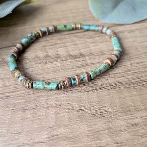 May include: A beaded bracelet featuring cylindrical turquoise-colored stone beads with dark matrix patterns, interspersed with small brown disc-shaped beads and metallic gold-toned spacers with a textured, beaded edge design.