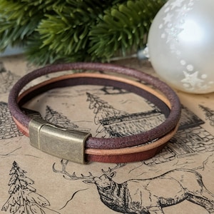 May include: A double-strand bracelet featuring one dark brown leather cord and one tan leather cord. The two strands are secured together by a rectangular, antique brass-toned metal magnetic clasp with a smooth, matte finish.