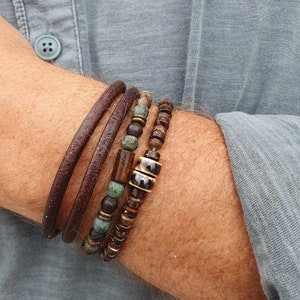 Może przedstawiać: Zestaw czterech bransoletek ułożonych na nadgarstku osoby. Bransoletki są wykonane ze skóry brązowej, drewna i zielonych i brązowych koralików.