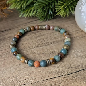 Puede incluir: Una pulsera de cuentas con piedras redondas alternas en tonos verde, marrón y naranja, intercaladas con pequeños espaciadores de metal de latón texturizado de estilo antiguo. Las cuentas están dispuestas en un patrón circular sobre un cordón.