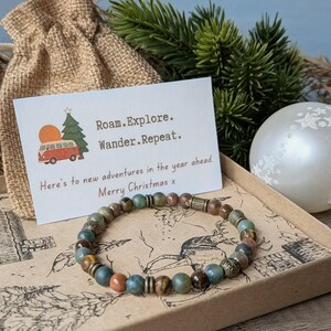 May include: A beaded bracelet featuring round stones in shades of green, brown, and tan, accented with metallic spacers. A white card displays the text Roam. Explore. Wander. Repeat. and Here's to new adventures in the year ahead. Merry Christmas x.