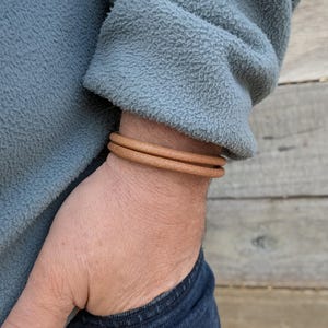 May include: A simple tan leather bracelet consisting of two thin, rounded cords that wrap around the wrist. The leather has a smooth, matte texture and a consistent light brown colour throughout the entire length of the double-layered band.