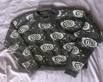 Könnte beinhalten: Ein grauer Strickpullover mit einem weißen Galaxie-Druck. Der Pullover hat einen Rundhalsausschnitt und lange Ärmel.