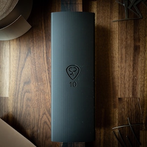May include: A dark gray rectangular object with the embossed logo "SP" inside a shield shape, and the number "10" below it. The object is on a wooden surface with other leather and metal objects.