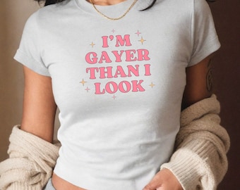 May include: A white t-shirt with a pink graphic that says "I'm Gayer Than I Look" with gold glitter stars around the text.