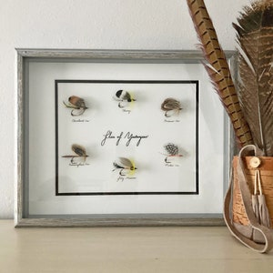 May include: A framed display titled Flies of Yesteryear featuring six individual fishing flies with feathers in various colors. Each fly is labeled with handwritten text including Cleveland Var, Cheney, Ondawa Var, Cunningham Var, Jock Scott, and Parker Var.