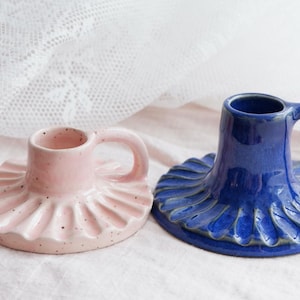 May include: Two ceramic candle holders, one pink with a ruffled base and one blue with a scalloped base. Both have a handle.