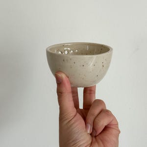 May include: A small, speckled, off-white ceramic bowl with a round shape and holes around the rim. The bowl is being held by a hand.