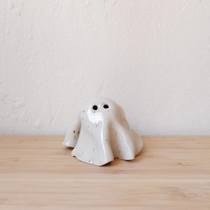 May include: A white ceramic ghost figurine with two black eyes. The ghost is sitting on a wooden surface.