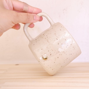 Peut inclure: Tasse en céramique crème avec un motif tacheté et un croissant de lune doré. La tasse a une grande anse et est tenue en biais, révélant sa forme. La tasse est posée sur une surface en bois clair.
