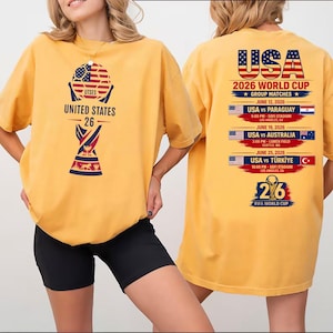 May include: Mustard yellow t-shirt featuring a graphic of a soccer ball with the American flag design and the text "UNITED STATES 26". The back of the shirt displays "USA 2026 WORLD CUP" with match details.