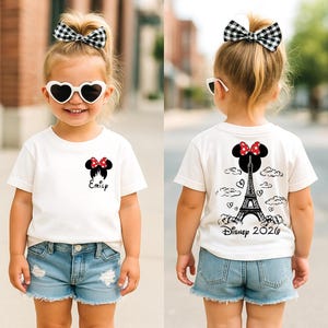 May include: A child wearing a white t-shirt with a Minnie Mouse design and the name "Emily" printed on the front. The back of the shirt features a Minnie Mouse design, the Eiffel Tower, and the text "Disney 2020". The child is also wearing denim shorts and a black and white checkered bow.