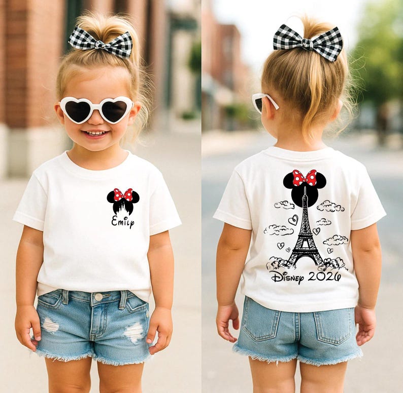 May include: A child wearing a white t-shirt with a Minnie Mouse design and the name "Emily" printed on the front. The back of the shirt features a Minnie Mouse design, the Eiffel Tower, and the text "Disney 2020". The child is also wearing denim shorts and a black and white checkered bow.