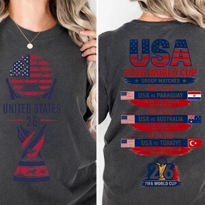 May include: Dark gray t-shirt with a graphic design featuring the American flag, the text "UNITED STATES 26", and a trophy. The back of the shirt displays "USA 2026 WORLD CUP" and match details.