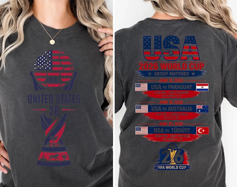 May include: Dark gray t-shirt with a graphic design featuring the American flag, the text "UNITED STATES 26", and a trophy. The back of the shirt displays "USA 2026 WORLD CUP" and match details.