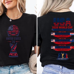 May include: Black t-shirt with a graphic design for the 2026 World Cup, featuring the USA flag and the text "UNITED STATES 26". The back of the shirt lists game dates and locations.