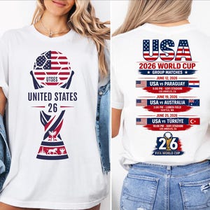 May include: White t-shirt featuring a soccer ball design with the American flag pattern and the text "UNITED STATES 26". The back of the shirt lists the USA 2026 World Cup group matches, including dates, times, and locations.