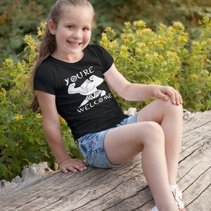 May include: A young person wearing a black t-shirt with a white graphic of a muscular figure and the text "You're Welcome". The person is sitting on a tree trunk and wearing denim shorts and white sandals.