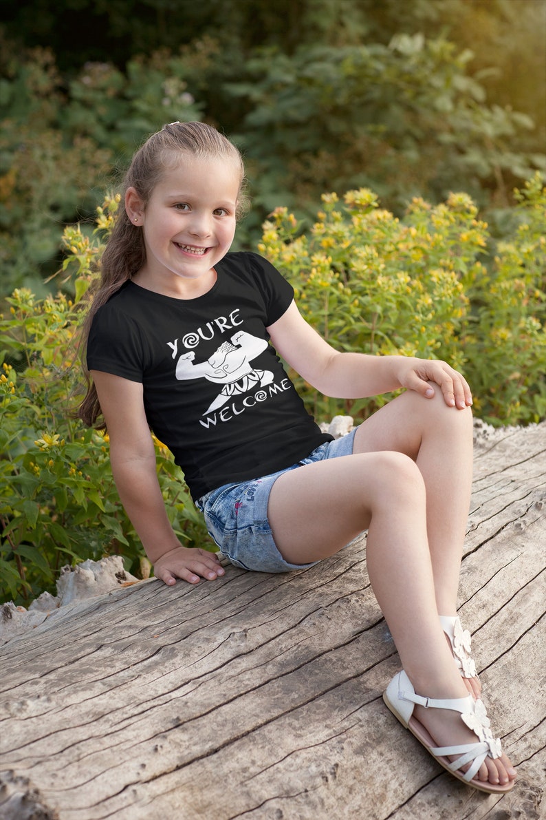 May include: A young person wearing a black t-shirt with a white graphic of a muscular figure and the text "You're Welcome". The person is sitting on a tree trunk and wearing denim shorts and white sandals.