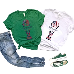 May include: Two t-shirts, one green and one white, with a graphic design featuring a soccer ball with an American flag design and the text "UNITED STATES 26". The image also includes a pair of blue jeans and green sneakers.