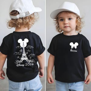 May include: Black t-shirt with a white Mickey Mouse silhouette and the name "Anton" printed below it. The back of the shirt features a design of the Eiffel Tower, clouds, hearts, and the text "Disney 2020". The child is wearing a white baseball cap.