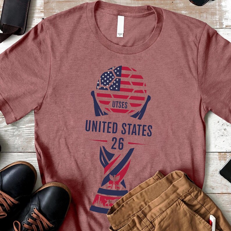 May include: A maroon t-shirt featuring a graphic of a soccer ball with the American flag design. The text "UTSES", "UNITED STATES", and "26" are printed below the ball in navy blue and red.