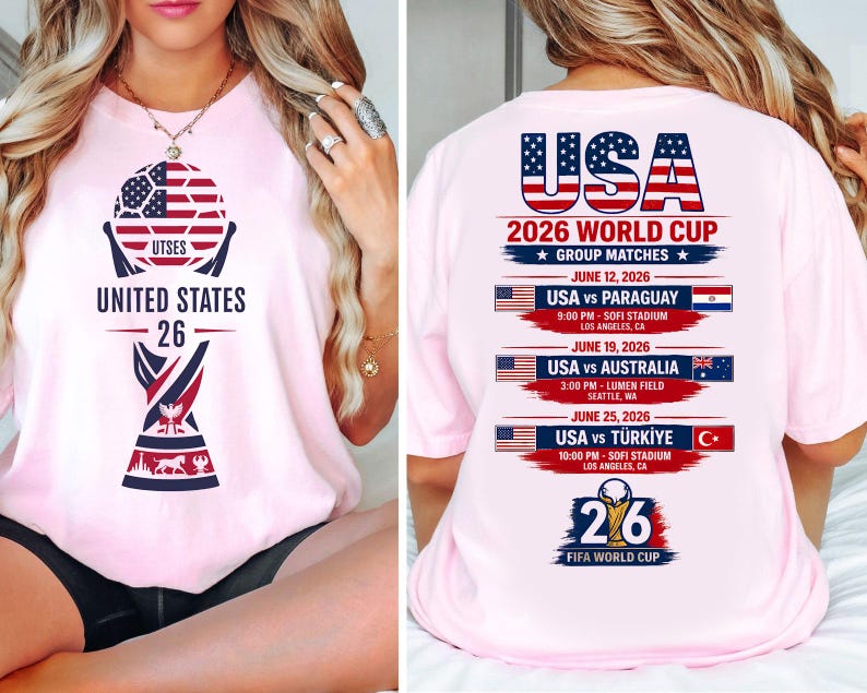 May include: Light pink t-shirt with a graphic featuring a soccer ball design with the text "UNITED STATES 26". The back of the shirt displays "USA 2026 WORLD CUP" with match details against Paraguay, Australia, and T&uuml;rkiye.