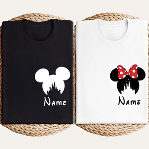 May include: Two t-shirts, one black and one white, each featuring a Disney-themed design. The black shirt has a white Mickey Mouse silhouette with a castle and the word "Name". The white shirt has a Minnie Mouse design with a red bow and the word "Name".