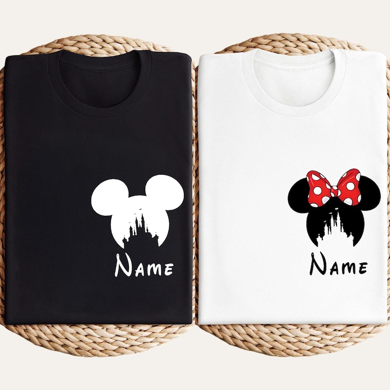 May include: Two t-shirts, one black and one white, each featuring a Disney-themed design. The black shirt has a white Mickey Mouse silhouette with a castle and the word "Name". The white shirt has a Minnie Mouse design with a red bow and the word "Name".