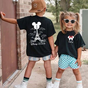 May include: Two children wearing black t-shirts with white graphics. One shirt features the Eiffel Tower and the text "Disney 2026". The other shirt has a Minnie Mouse design and the name "Emily". They are wearing shorts, socks, and sneakers.