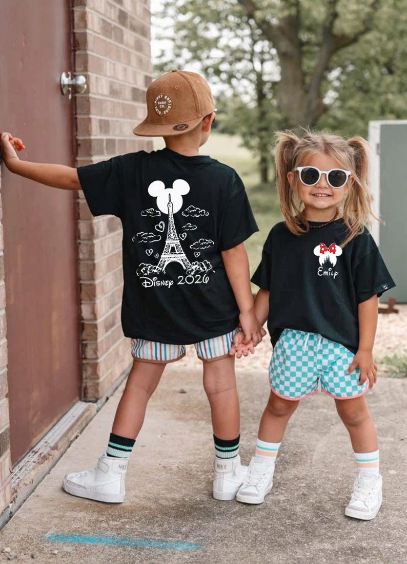 May include: Two children wearing black t-shirts with white graphics. One shirt features the Eiffel Tower and the text "Disney 2026". The other shirt has a Minnie Mouse design and the name "Emily". They are wearing shorts, socks, and sneakers.