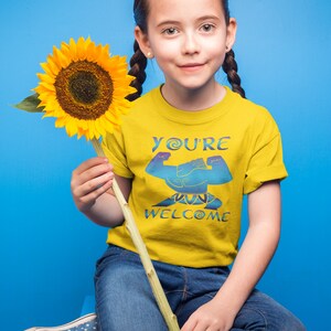 May include: A young person wearing a yellow t-shirt with the text "You're Welcome" and a blue graphic. They are holding a sunflower and wearing blue jeans.