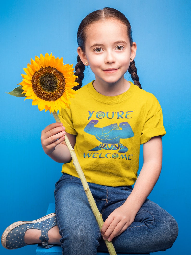 May include: A young person wearing a yellow t-shirt with the text "You're Welcome" and a blue graphic. They are holding a sunflower and wearing blue jeans.