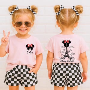 May include: A young child wearing a pink t-shirt and black and white checkered shorts. The shirt features a Minnie Mouse design with the name "Emily" printed below. The back of the shirt has a design of the Eiffel Tower and the text "Disney 2026".