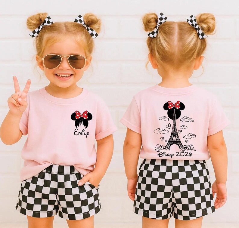 May include: A young child wearing a pink t-shirt and black and white checkered shorts. The shirt features a Minnie Mouse design with the name "Emily" printed below. The back of the shirt has a design of the Eiffel Tower and the text "Disney 2026".