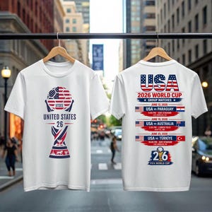 May include: White t-shirts with a soccer-themed design. One shirt features a soccer ball graphic with the text "UNITED STATES 26". The other shirt displays "USA 2026 WORLD CUP" with match details, including dates, opponents, and locations.